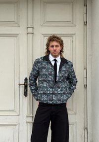 Veste à motifs avec un design en vitrail aux couleurs sombres, associée à une chemise blanche et une cravate noire, portée avec un pantalon noir.
