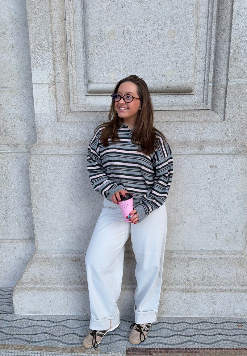 Young woman wearing glasses, striped long-sleeve shirt, and white pants, smiling and holding a pink coffee cup against stone wall background.