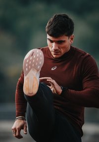 Athlète portant un haut moulant bordeaux à manches longues et un pantalon noir, exécutant un coup de pied haut, mettant en valeur une chaussure de course de couleur claire avec une semelle texturée.