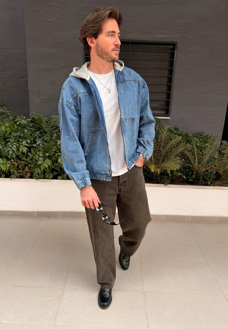 Homme portant une veste en jean bleue sur une chemise blanche, pantalon foncé, chaussures noires, tenant des lunettes de soleil, marchant près des plantes et d'un mur gris.