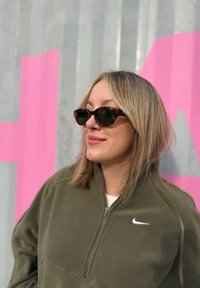 Smeđe kornjačaste sunčane naočale, maslinasto zelena zip-up flis jakna s bijelim Nike logotipom, svijetla kosa i meka tekstura na ružičastoj pozadini.