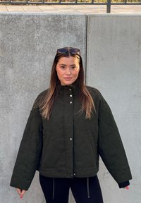 Črno prešito jakno z visokim ovratnikom, sprednjimi gumbi na sponko in velikimi žepi. Ima ohlapen kroj in teksturirano tkanino.