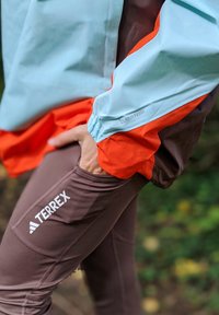 Terrex outdoor jakke har lys blå og orange farveblokke, mens brune legginger viser et logo og funktionelt lomme-design.