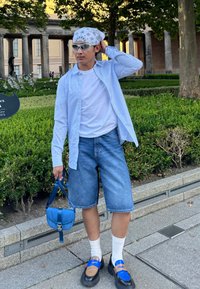Camicia oversize a righe blu chiaro sopra una t-shirt bianca, abbinata a pantaloni shorts di jeans. Gli accessori includono un bandana a fantasia e una piccola borsa blu.
