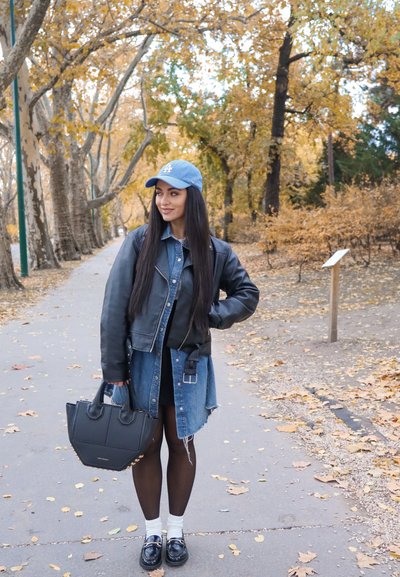 Chaqueta de cuero negra, vestido vaquero, gorra de béisbol azul, bolso negro, calcetines blancos, mocasines negros; de pie sobre un camino cubierto de hojas.