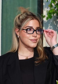 Czarne okulary w ramkach z niebieskimi akcentami, noszone przez osobę z długimi, prostymi, blond włosami, szarym topem i czarnym żakietem. Widać srebrną bransoletkę.