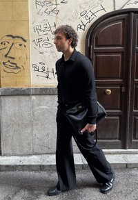 Homme en tenue noire marchant dans la rue, tenant une grande pochette noire, avec des graffitis sur le mur et une porte en bois sombre derrière lui.