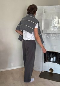 Suéter a rayas grises sobre los hombros, camiseta blanca, pantalones grises, de pie frente a una chimenea de azulejos blancos con velas.