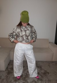 Veste courte à motif camouflage, pantalon large blanc brillant, et bonnet rayé vert vif. Chaussettes roses éclatantes visibles.