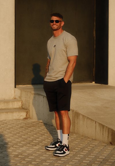 Camiseta gris de Nike, pantalones cortos negros y zapatillas negras con acentos rojos. De pie en unos escalones de cemento, usando gafas de sol y calcetines blancos.