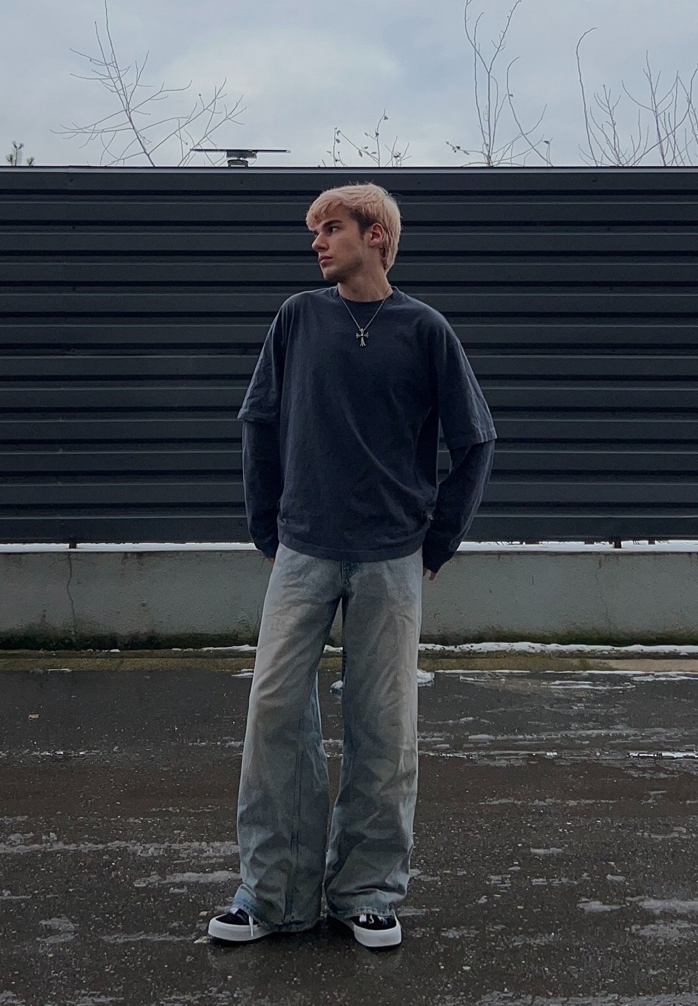 Jeune personne aux cheveux blonds debout sur un trottoir mouillé, portant une chemise à manches longues superposée foncée, un jean large clair, des baskets et un collier avec une croix.