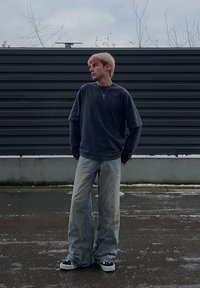 Jeune personne aux cheveux blonds debout sur un trottoir mouillé, portant une chemise à manches longues superposée foncée, un jean large clair, des baskets et un collier avec une croix.