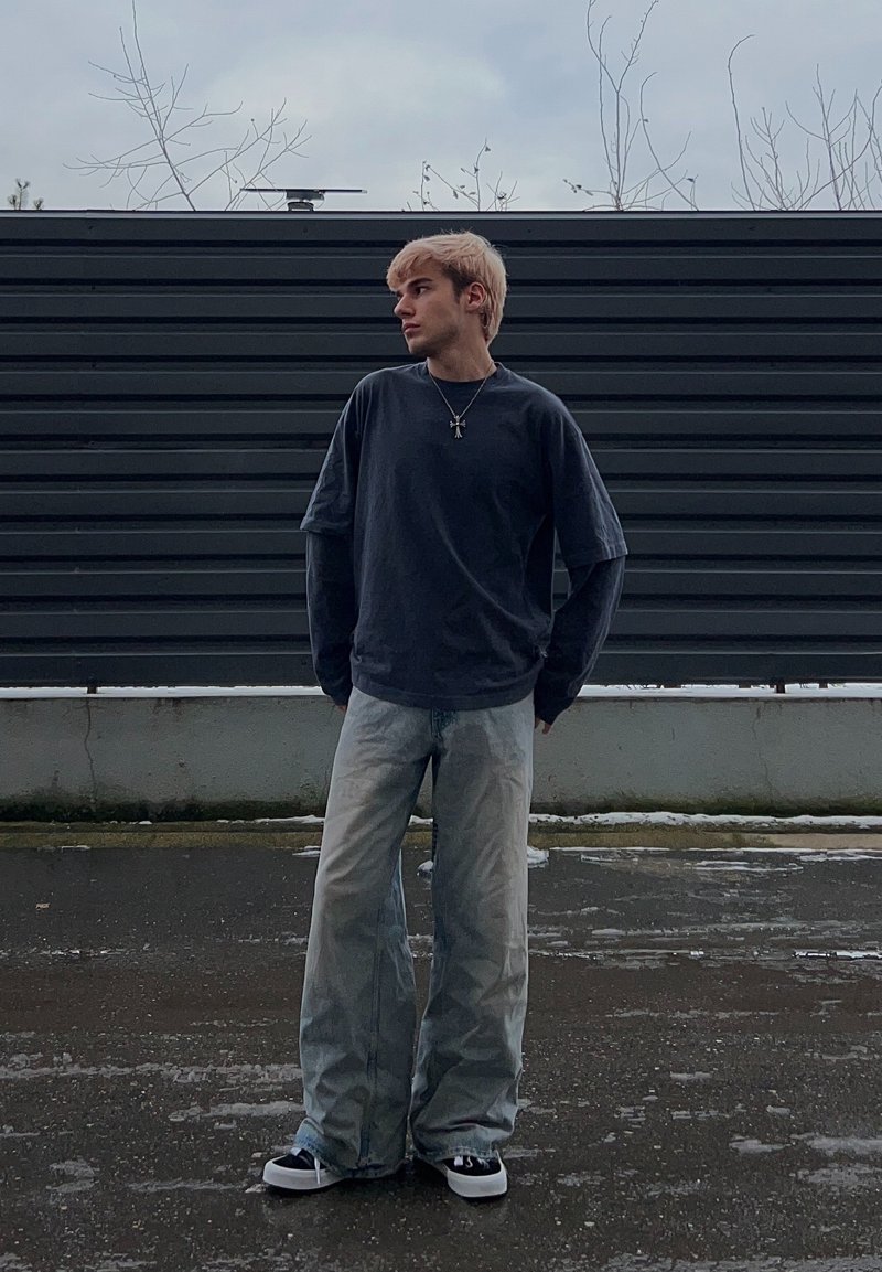 Une jeune personne aux cheveux blonds se tient sur un trottoir mouillé, portant une chemise à manches longues sombre en superposition, un large jean clair, des baskets et un collier croix.