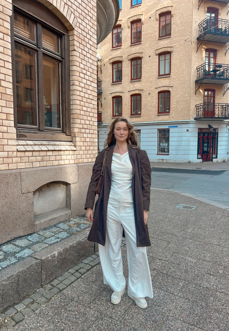 Lichte bruine jas over een witte top enwijde broek. Sneakers met een gemêleerde zool. Baksteen- en stenen gebouw op de achtergrond.