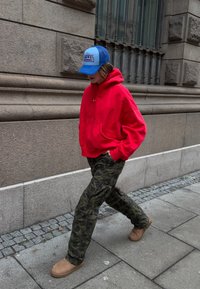 Sudadera roja, pantalones de camuflaje y botas beige. Gorra de béisbol azul y blanca con logo. Atuendo casual de estilo urbano. Entorno urbano.