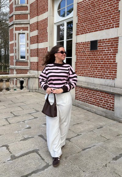 Joven mujer con gafas de sol oscuras y suéter de rayas caminando sobre pavimento de piedra junto a un edificio de ladrillo rojo, sosteniendo un bolso marrón.