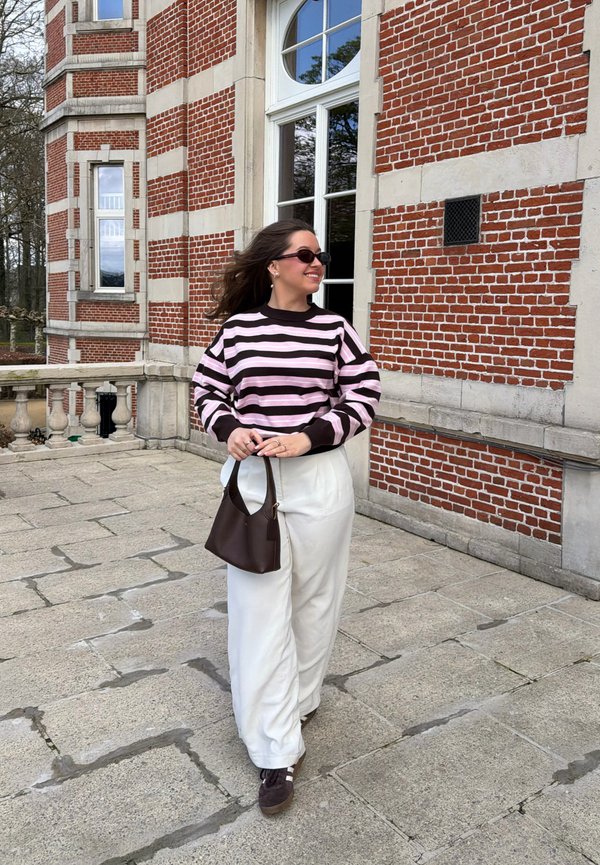 Jeune femme avec des lunettes de soleil foncées et un pull rayé marchant sur un pavement en pierre à côté d'un bâtiment en brique rouge, tenant un sac à main marron.