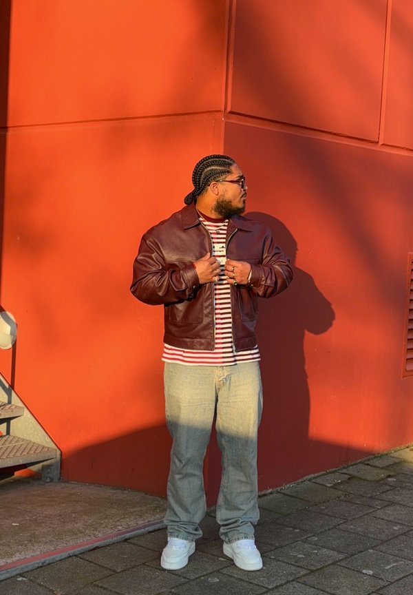 Man with braided hair wearing glasses, maroon leather jacket, striped shirt, light jeans, and white sneakers standing against an orange wall.