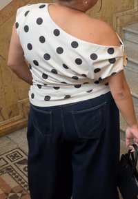 Γυναίκα που φοράει λευκό τοπ με μαύρες πουά, σκούρο μπλε παντελόνι, κρατώντας μια μαύρη τσάντα, στέκεται κοντά σε δάπεδο με πλακάκια με σχέδιο και σκάλες.