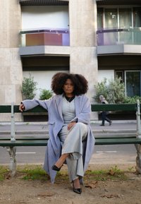 Γκρι μάλλινο παλτό πάνω από γκρι τοπ και ανοιχτόχρωμο παντελόνι. Μαύρα κοφτά παπούτσια, σγουρά μαλλιά, καθισμένος σε μια πράσινη παγκάκι, αστικό φόντο.