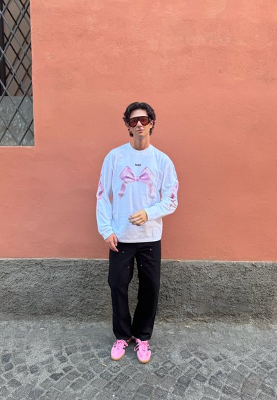 Camisa blanca de manga larga con gráficos de moño rosa, pantalones negros y zapatillas rosas. El fondo presenta una pared de color coral con una ventana.