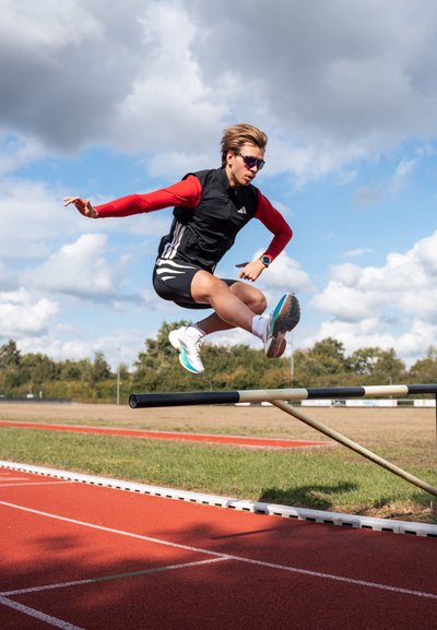 Atleta con chaleco negro y mangas rojas, y pantalones cortos negros, salta sobre una barra horizontal en una pista. Zapatillas de correr color azul claro, al aire libre.