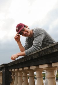 Casquette bordeaux avec un "P", pull gris en maille torsadée, appuyé contre une rambarde en pierre sous un ciel nuageux. Paysage naturel en arrière-plan.