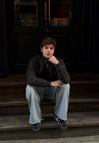 Fiatal férfi ül fa lépcsőn, fekete cipzáras dzsekit és világoskék farmert visel, sötét tornacipővel. A háttérben egy ajtó látható.