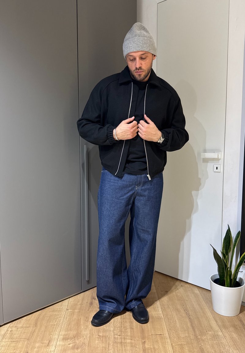 Veste bomber noire zippée portée sur un t-shirt noir, associée à un jean denim bleu large et des chaussures noires, complétée par un bonnet gris.