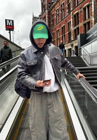 Black leather jacket over a white hoodie, grey cargo trousers. Green cap with a logo, holding a smartphone on an escalator. Urban background.