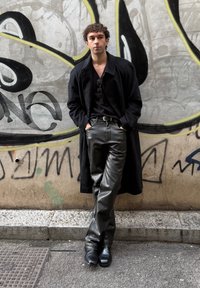 Jeune homme portant un long manteau noir, une chemise noire, un pantalon en cuir et des bottes, debout les mains dans les poches contre un mur couvert de graffitis.