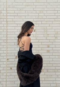 Femme aux cheveux foncés, portant une robe noire à bretelles, tenant un manteau noir bordé de fourrure, debout de profil contre un mur en briques gris clair.