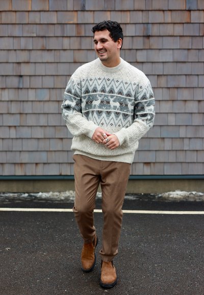 Hombre caminando al aire libre con un suéter blanco y gris con patrón de renos, pantalones marrones y botas de ante marrones, sonriendo y mirando hacia un lado.