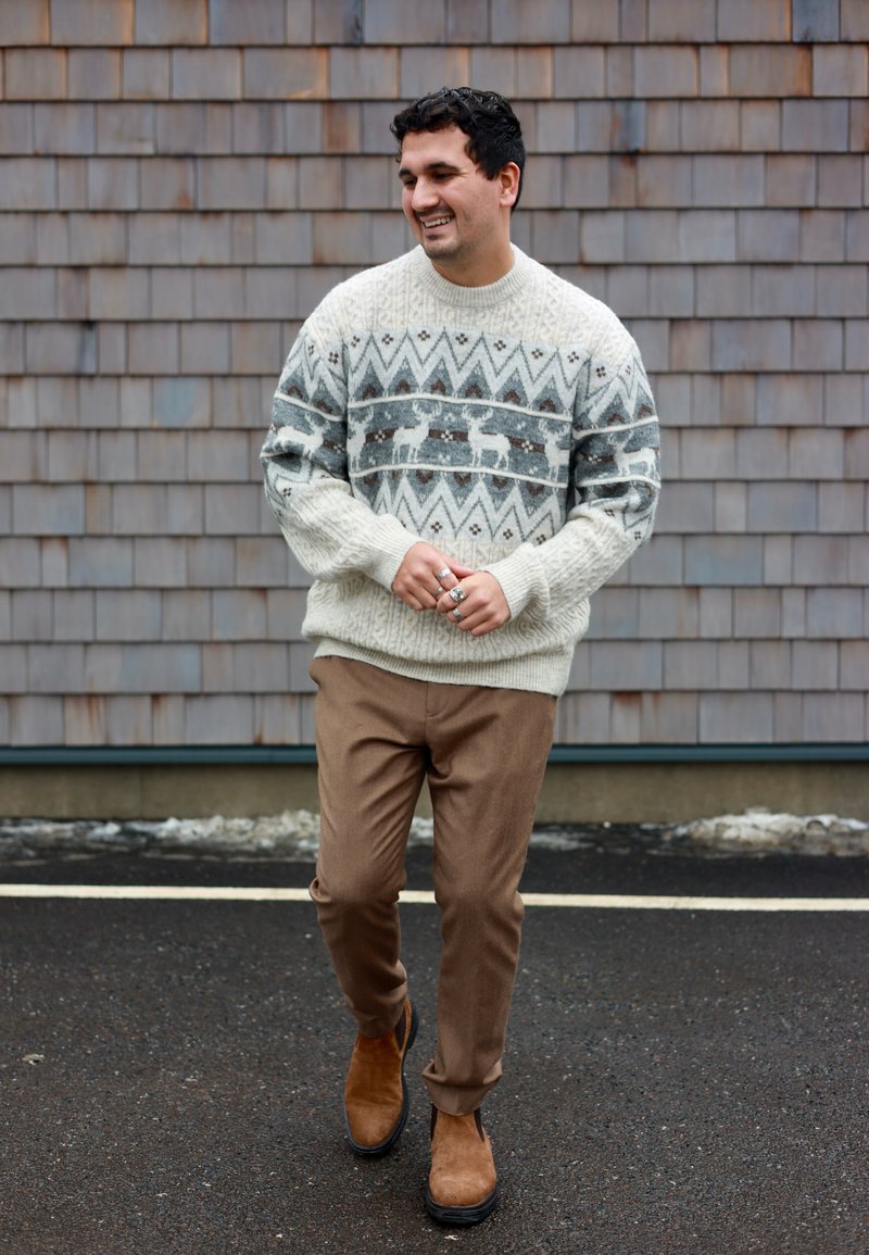 Mann, der im Freien geht und einen weißen und grauen Pullover mit Rentiermuster, braune Hosen und braune Wildlederstiefel trägt, lächelt und zur Seite schaut.