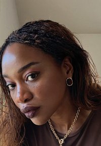 Femme aux cheveux tressés et lâchés portant un rouge à lèvres foncé, des boucles d'oreilles en or en forme de cercle et un collier chaîne épais en or, regardant légèrement vers la gauche.