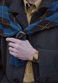 Homme portant un manteau marron et une chemise jaune ajuste une écharpe bleue et jaune à motifs, montrant une montre numérique dorée au poignet gauche.
