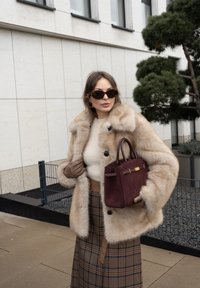 Cappotto in pelliccia sintetica color cammello, maglione in lana color crema, gonna a quadri marrone. Porta una borsa burgundy con dettagli dorati e indossa guanti marroni.