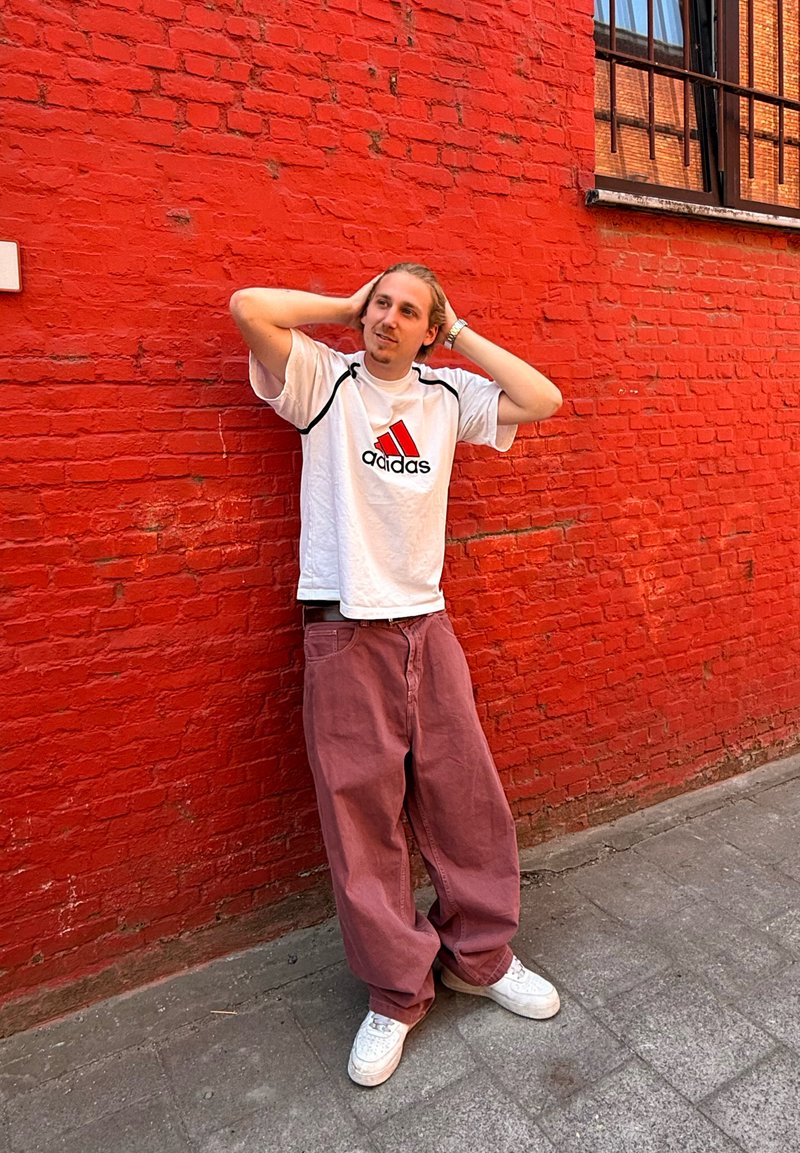 Hvid T-shirt med sorte og røde accenter og Adidas-logo, parret med oversized pink bukser og hvide sneakers. Rød murstensvæg i baggrunden.