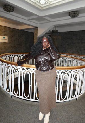 Mujer con cabello largo y rizado apoya en una barandilla blanca dentro de un edificio, vistiendo una chaqueta de cuero oscura, falda marrón y botas blancas.