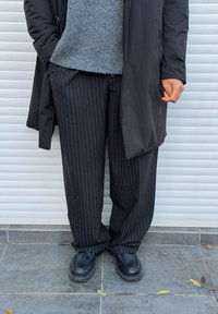 Pantalones negros con rayas finas y un corte holgado, combinados con un suéter gris texturizado y zapatos negros sin cordones. Se ve parcialmente un abrigo negro liso.