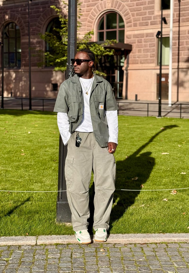 Homme portant des lunettes de soleil, une chemise verte superposée à une chemise blanche et un pantalon cargo, appuyé contre un poteau dans un parc ensoleillé avec un chemin en pavés.