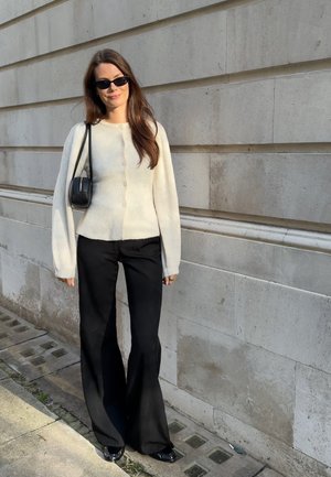 Mujer con pantalones negros de pierna ancha, suéter crema abotonado, bolso negro de hombro y gafas de sol, de pie contra una pared de piedra beige al aire libre.
