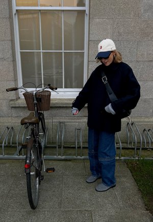 Persona con abrigo oscuro y gorra junto a una bicicleta estacionada con una cesta frontal, al lado de un edificio con una ventana grande.