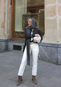 Zwarte cardigan over een witte top, witte jeans, grijze sjaal en taupe enkellaarzen. Houdt een crème-kleurige, gestructureerde handtas vast. Stedelijke omgeving.