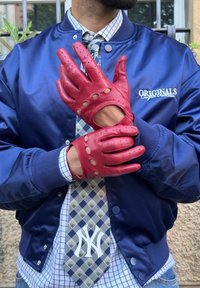 Guanti in pelle rossa con dettagli perforati e sezioni ritagliate, indossati da una persona in giacca blu e cravatta a fantasia.