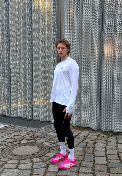 Joven con camiseta blanca de manga larga, mallas negras, calcetines blancos y zapatillas deportivas fucsias brillantes, de pie sobre adoquines junto a una pared metálica texturizada.