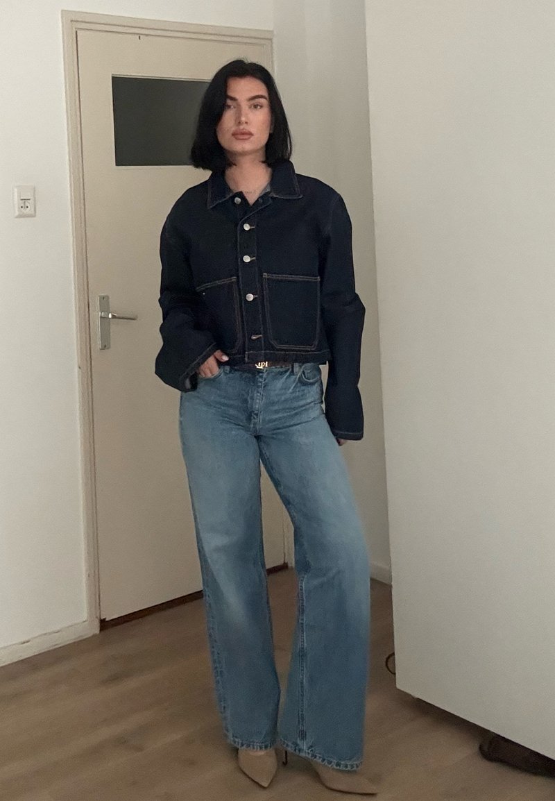 Personne debout à l'intérieur portant une veste en jean foncé, un jean large bleu clair, et des talons pointus beiges.