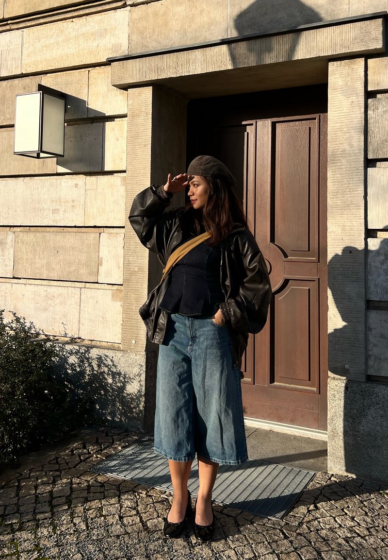 Femme portant un béret, une veste en cuir et un pantalon large, se protégeant les yeux du soleil, debout près d'une porte en bois sur une rue pavée de pierres.