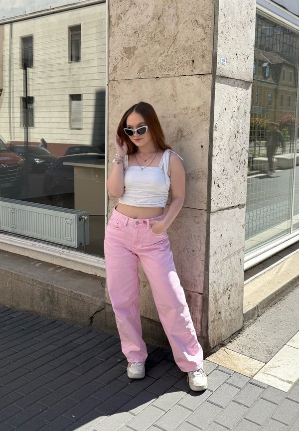 Jeune femme en haut blanc, pantalon rose et baskets blanches, debout sur le trottoir contre un mur en pierre beige, portant des lunettes de soleil cat-eye.