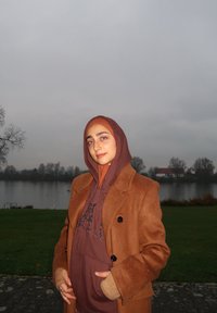 Brun dobbeltradet frakke over en brun hættetrøje; matchende hijab; udendørs ved en sø; overskyet himmel; lette dråber på frakken.
